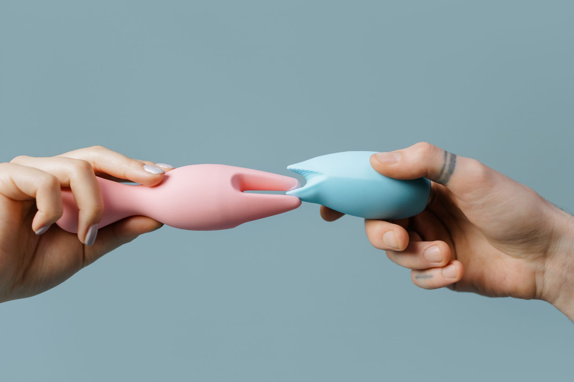 Hands holding colorful silicone clitoral vibrators against a neutral background